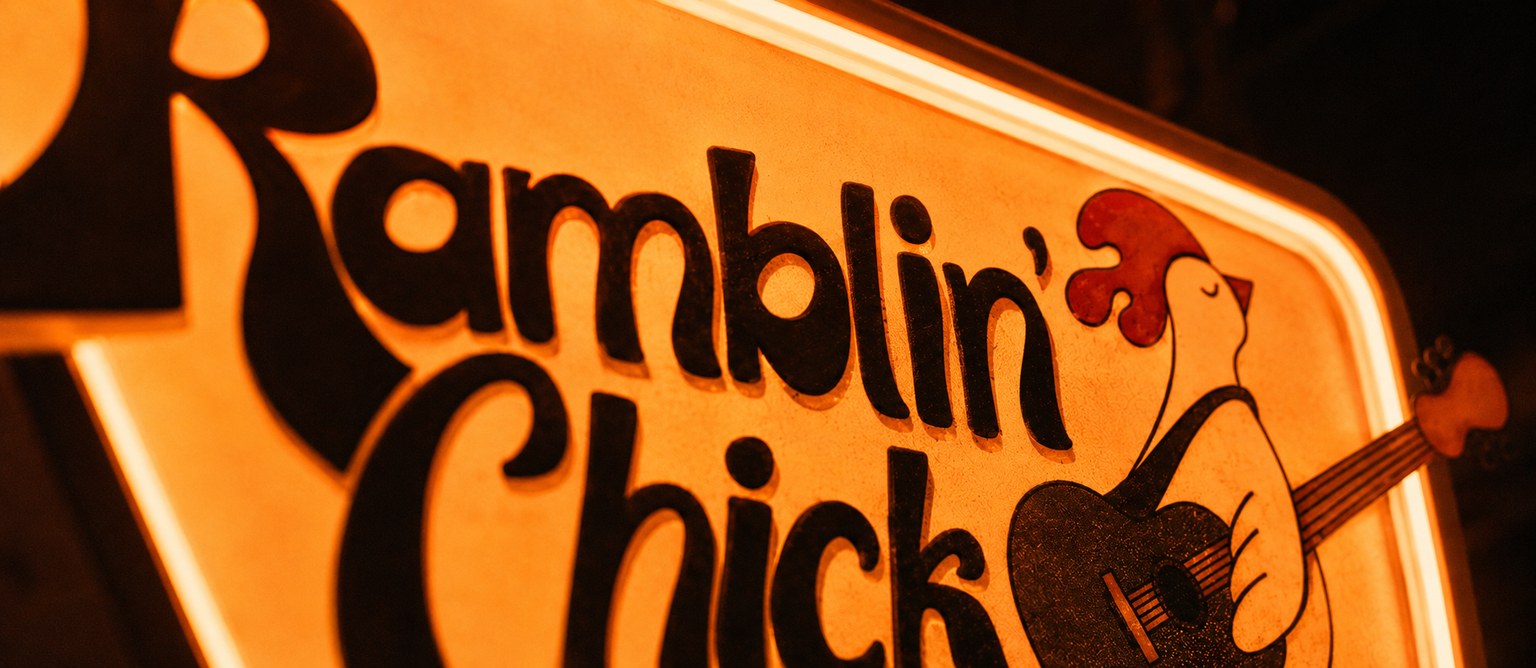 Warm orange illuminated Ramblin’ Chick sign with cropped lettering and a guitar-playing chicken.